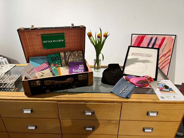 Trauerbücher und Beispiele der Erinnerungswerkstatt Offener Koffer mit Büchern auf einem Holztisch neben einer Vase mit Tulpen, und Beispiele wie Kissen , Wärmflaschenbezug