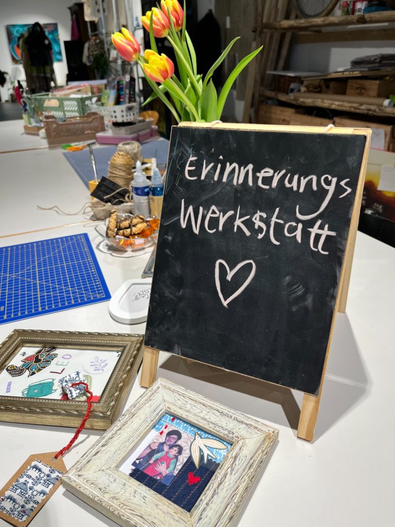 Willkommen in der Errinnerungswerkstatt Kreidetafel mit der Aufschrift 'Erinnerungs Werkstatt' und einem Herz, umgeben von Tulpen, gerahmten Fotos und Bastelmaterialien auf einem T